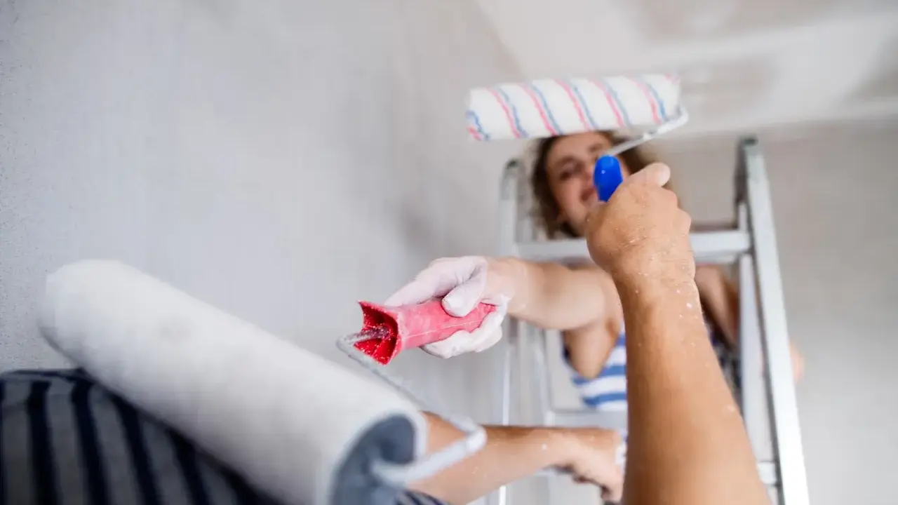 Dwie osoby malują sufit. Jedna stoi na drabinie, druga podaje jej wałek. Trwa nakładanie akrylu przy suficie.