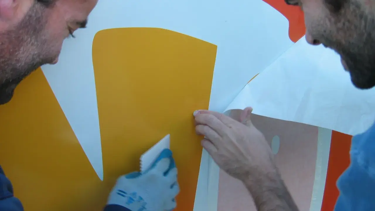 Dwóch mężczyzn pokazuje, jak nakleić naklejkę na ścianę. Jeden wygładza ją raklem, drugi odkleja papier.