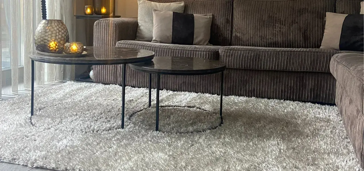 Miękki dywan w kolorze greige otula salon. Na nim dwa stoliki kawowe z marmurowymi blatami i aksamitna kanapa.