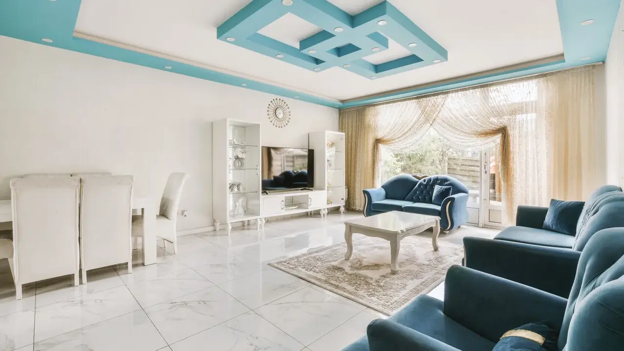 Nowoczesny salon z błękitnym, geometrycznym, dekoracyjnym malowidłem na suficie. Wygodne, granatowe sofy i białe meble tworzą elegancką przestrzeń.