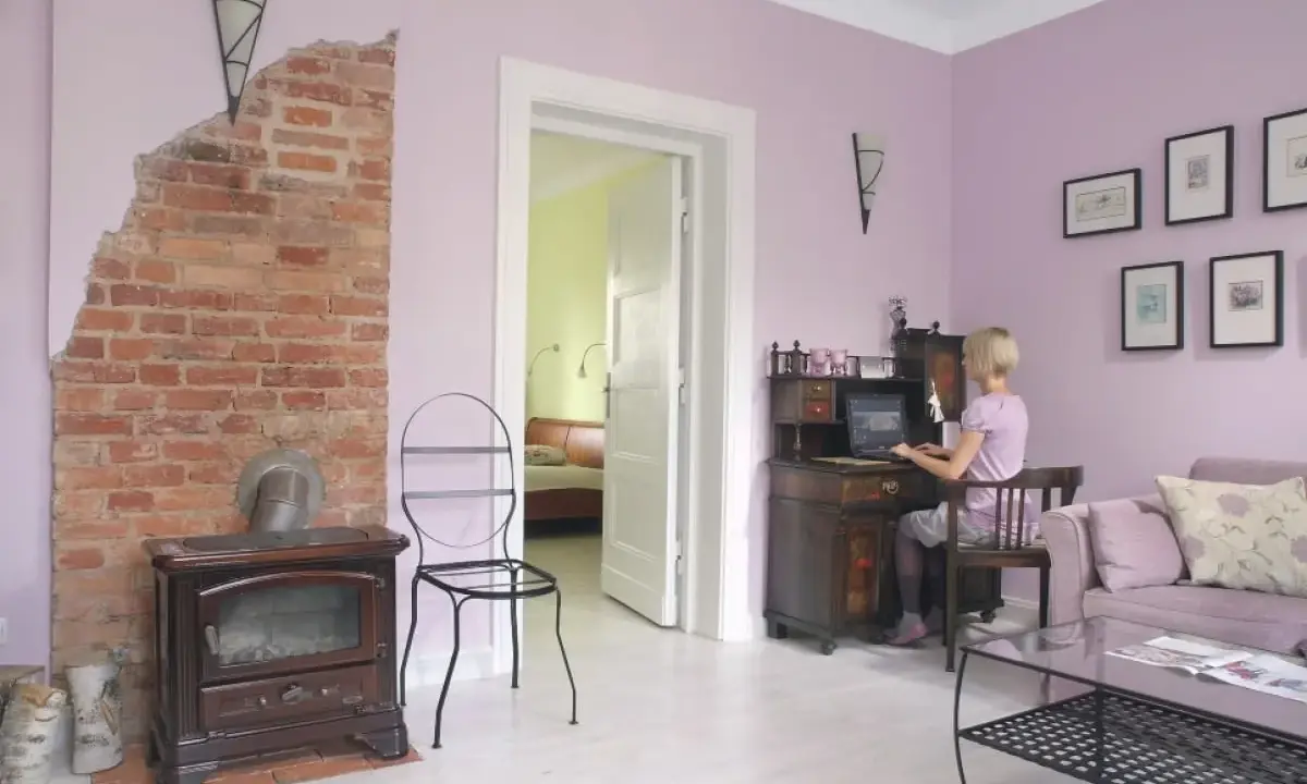 Pokój z ceglanym piecem i biurkiem, gdzie pracuje kobieta. Ściany w odcieniu **kolor lawendowy**.