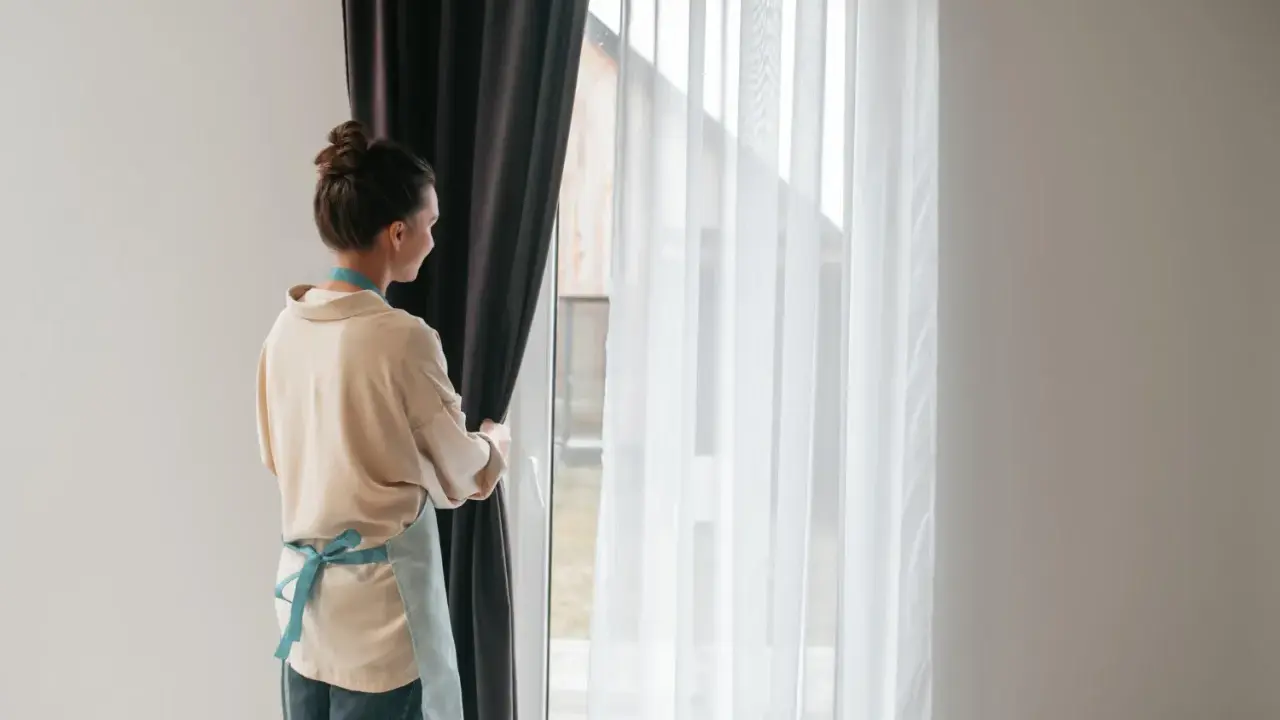Kobieta w fartuszku zasłania okno ciemną zasłoną. Widać białe firanki i karnisz z wieloma ząbkami.