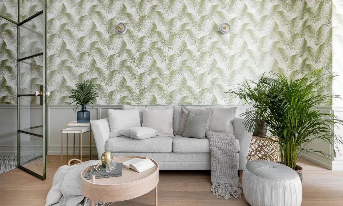Jasne, zielone tapety z motywem liści paproci tworzą tropikalny klimat w salonie. Sofa i stolik kawowy dopełniają przytulną aranżację.