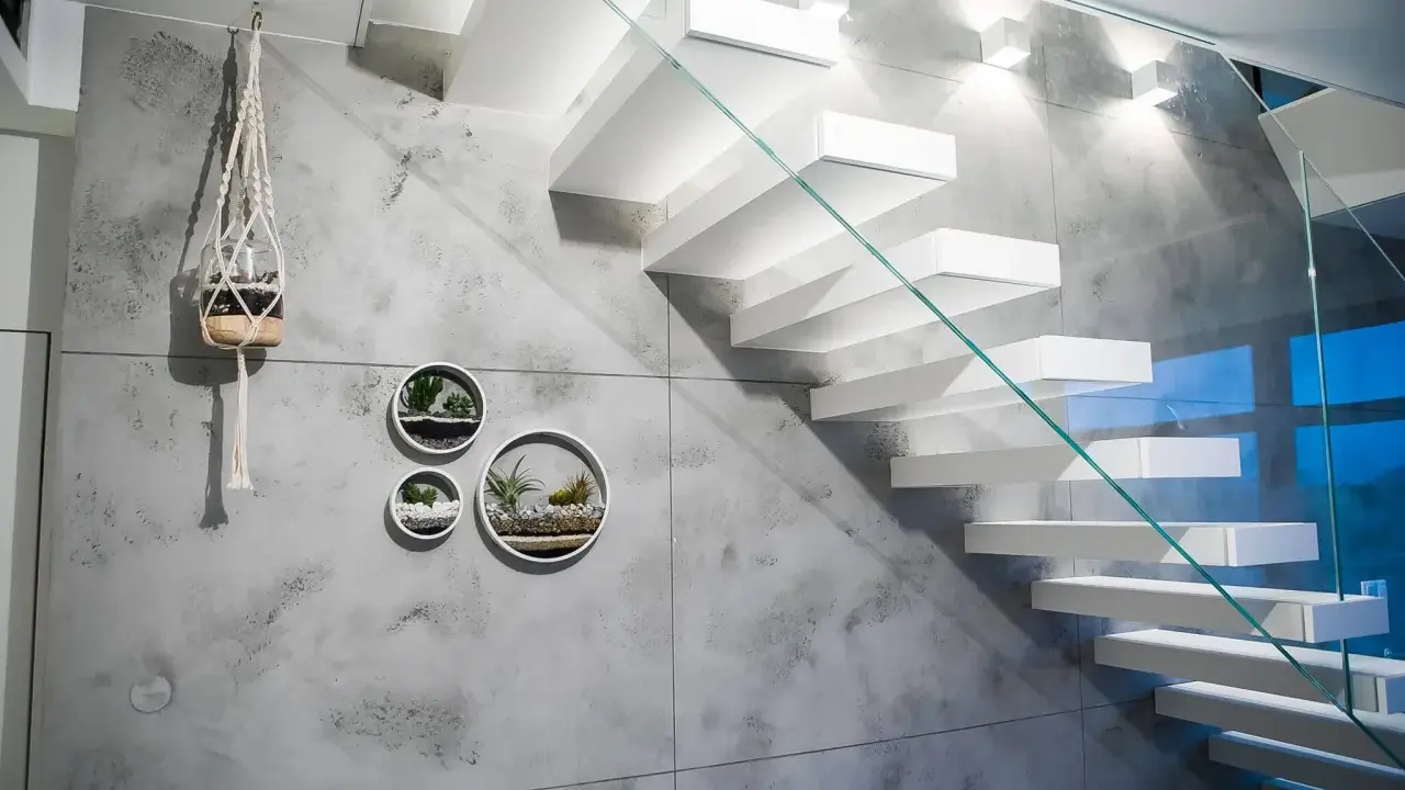 Nowoczesna klatka schodowa z białymi, lewitującymi stopniami i szklaną balustradą. Ściana ozdobiona betonem architektonicznym i wiszącymi doniczkami z sukulentami.
