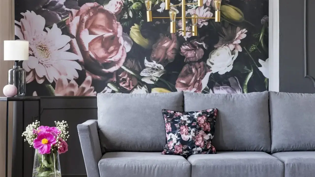 Szara sofa z poduszką w kwiaty, na tle bogatej fototapety. Idealna, by stworzyć przytulną atmosferę.