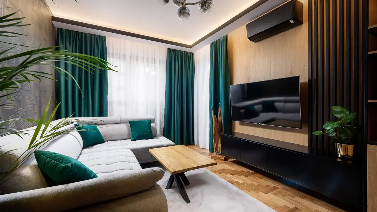 Nowoczesny salon z szarą kanapą i zielonymi poduszkami. Po prawej stronie ściana ozdobiona pionowymi lamelami w narożniku, na niej telewizor.