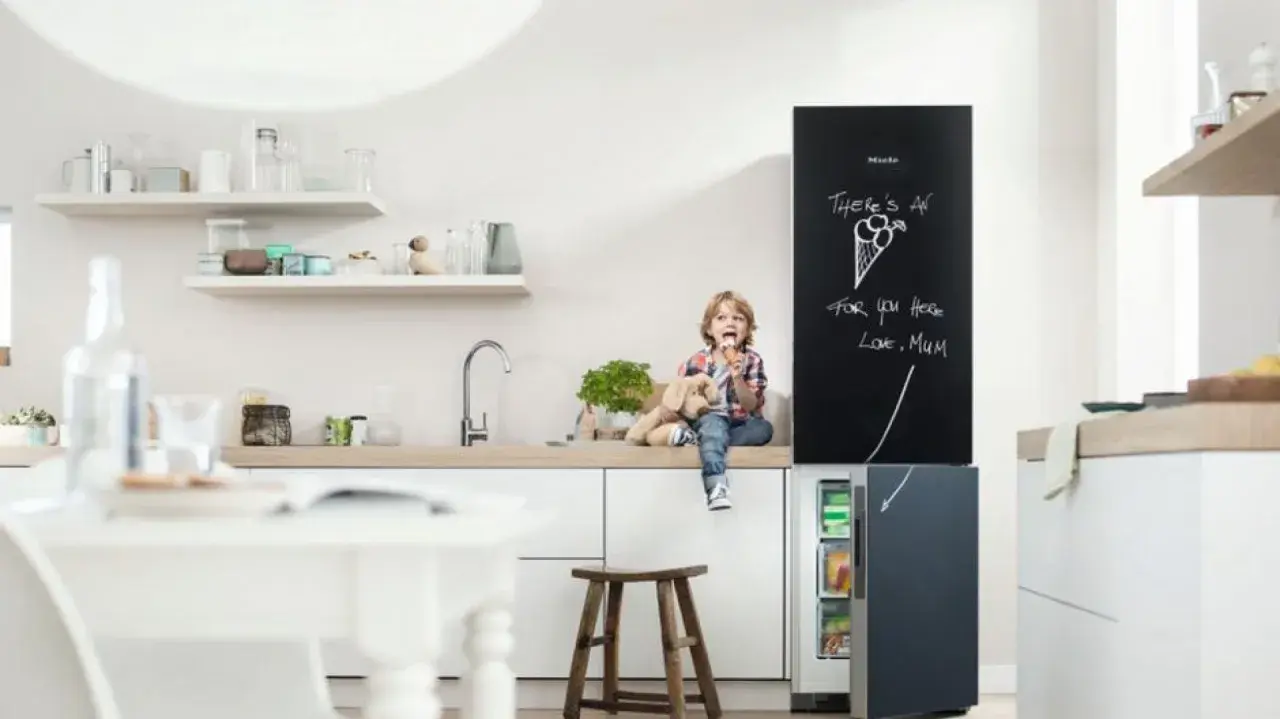 Chłopiec z pluszowym psem siedzi na blacie w kuchni, jedząc lody. Obok lodówka z tablicą, na której mama napisała "There's an ice cream for you here". Inspiracje na tablicę w kuchni.