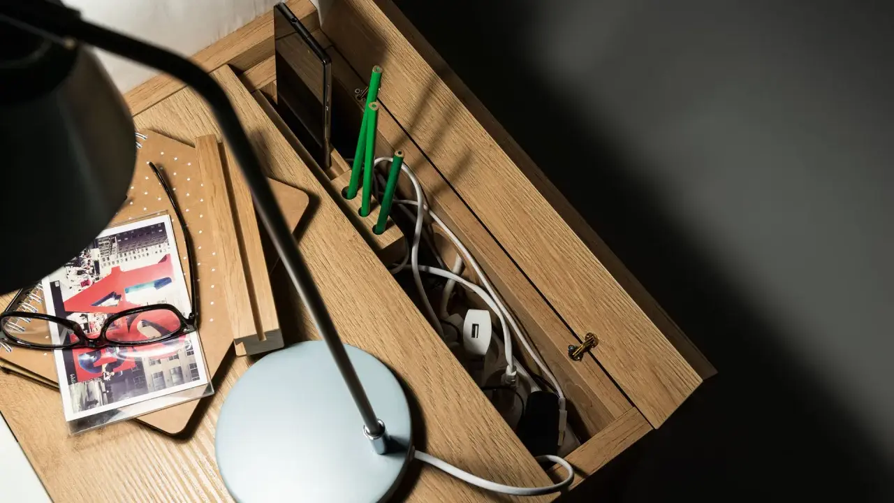Drewniana szafka nocna z przegródkami, gdzie można schować wystające kable ze ściany. Na blacie lampka, okulary i notatnik.