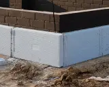 Białe, ekstrudowane bloki styropianowe tworzą fundament domu, otaczając ciemną ceglaną ścianę.