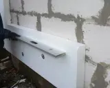 Ręka w rękawicy przykłada poziomicę do styropianu przy ścianie. To pokazuje, czym szlifować styropian, by uzyskać równą powierzchnię.