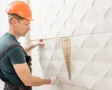 Budowlaniec w kasku montuje białe płytki gipsowe na ścianę, tworząc nowoczesne aranżacje.