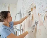 Kobieta w rękawiczkach zeskrobuje starą tapetę. Zastanawia się, czym podkleić nową, by dobrze trzymała się ściany.