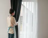 Kobieta w fartuszku zasłania okno ciemną zasłoną. Widać białe firanki i karnisz z wieloma ząbkami.