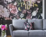 Szara sofa z poduszką w kwiaty, na tle bogatej fototapety. Idealna, by stworzyć przytulną atmosferę.