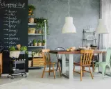 Tablica z listą zakupów nad stołem i krzesłami w kuchni. Widać na niej napisy kredą: "cheese", "milk", "bread", "joghurt", "dipes", "lemon", "wine", "banana", "olive", "tomato".