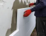 Pracownik w czerwonych rękawicach układa płyty styropianowe, rozpoczynając naprawę elewacji styropianowej.