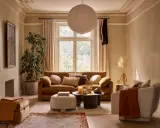 Przytulny salon z kanapą w kolorze karmelowym, ozdobioną poduszkami. Obok fotel, puf i stolik kawowy.