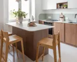 Jasna kuchnia w stylu skandynawskim z wyspą. Drewniane fronty szafek, jasny blat i minimalistyczne hokery tworzą przytulną atmosferę.
