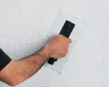 Ręka z kielnią wygładza tynk na styrodurze. Dowiedz się, jak wykończyć styrodur, by uzyskać gładką ścianę.