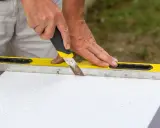 Ręce trzymają nóż, którym cięty jest styropian wzdłuż żółtej poziomicy.