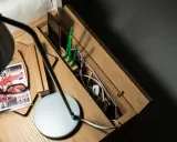 Drewniana szafka nocna z przegródkami, gdzie można schować wystające kable ze ściany. Na blacie lampka, okulary i notatnik.