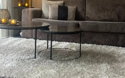 Miękki dywan w kolorze greige otula salon. Na nim dwa stoliki kawowe z marmurowymi blatami i aksamitna kanapa.