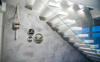 Nowoczesna klatka schodowa z białymi, lewitującymi stopniami i szklaną balustradą. Ściana ozdobiona betonem architektonicznym i wiszącymi doniczkami z sukulentami.