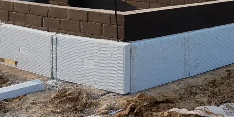 Białe, ekstrudowane bloki styropianowe tworzą fundament domu, otaczając ciemną ceglaną ścianę.