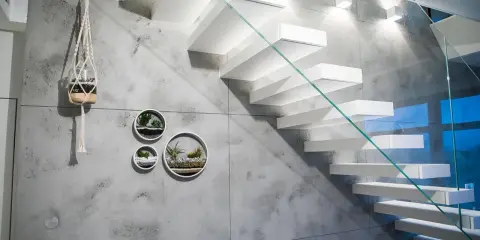 Nowoczesna klatka schodowa z białymi, lewitującymi stopniami i szklaną balustradą. Ściana ozdobiona betonem architektonicznym i wiszącymi doniczkami z sukulentami.