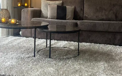 Miękki dywan w kolorze greige otula salon. Na nim dwa stoliki kawowe z marmurowymi blatami i aksamitna kanapa.