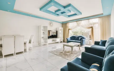 Nowoczesny salon z błękitnym, geometrycznym, dekoracyjnym malowidłem na suficie. Wygodne, granatowe sofy i białe meble tworzą elegancką przestrzeń.
