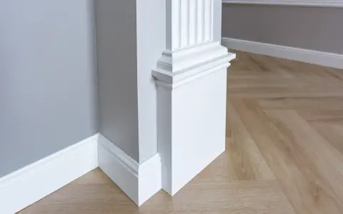 Biały pilaster z żłobieniami i elegancką bazą, idealny do wnętrzarskiego bloga. Drewniana podłoga w jodełkę i szara ściana tworzą stylowe tło.