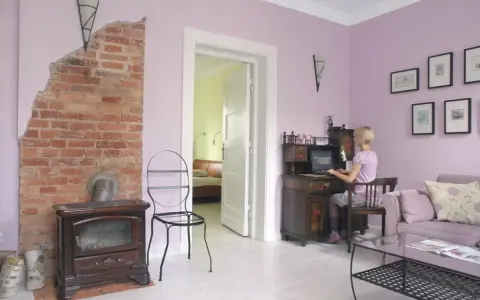 Pokój z ceglanym piecem i biurkiem, gdzie pracuje kobieta. Ściany w odcieniu **kolor lawendowy**.