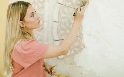 Młoda kobieta z piegami i blond włosami maluje pędzlem ozdobną sztukaterię na ścianie.