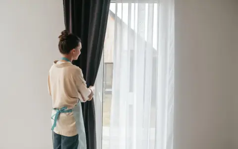 Kobieta w fartuszku zasłania okno ciemną zasłoną. Widać białe firanki i karnisz z wieloma ząbkami.
