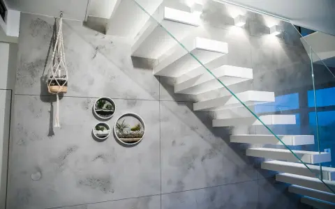 Nowoczesna klatka schodowa z białymi, lewitującymi stopniami i szklaną balustradą. Ściana ozdobiona betonem architektonicznym i wiszącymi doniczkami z sukulentami.