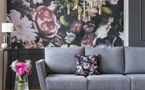 Szara sofa z poduszką w kwiaty, na tle bogatej fototapety. Idealna, by stworzyć przytulną atmosferę.