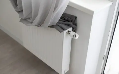 Biały grzejnik z termostatem, na którym leży szara zasłona. Widać, że odległość firanki od grzejnika jest niewielka, co może wpływać na cyrkulację powietrza.