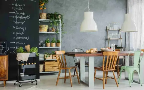 Tablica z listą zakupów nad stołem i krzesłami w kuchni. Widać na niej napisy kredą: "cheese", "milk", "bread", "joghurt", "dipes", "lemon", "wine", "banana", "olive", "tomato".