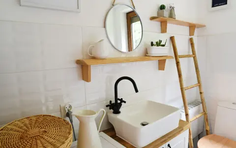 Urocza, mała łazienka w stylu retro z białymi kafelkami, drewnianymi półkami, okrągłym lustrem i bambusową drabiną.