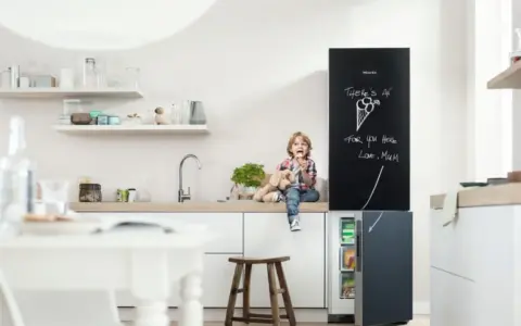 Chłopiec z pluszowym psem siedzi na blacie w kuchni, jedząc lody. Obok lodówka z tablicą, na której mama napisała "There's an ice cream for you here". Inspiracje na tablicę w kuchni.