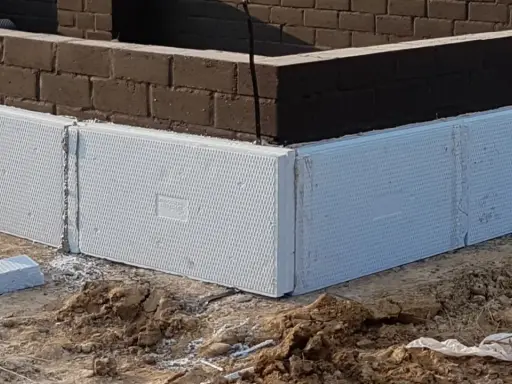 Białe, ekstrudowane bloki styropianowe tworzą fundament domu, otaczając ciemną ceglaną ścianę.