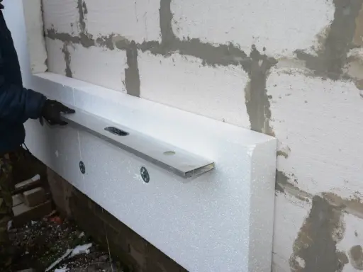 Ręka w rękawicy przykłada poziomicę do styropianu przy ścianie. To pokazuje, czym szlifować styropian, by uzyskać równą powierzchnię.