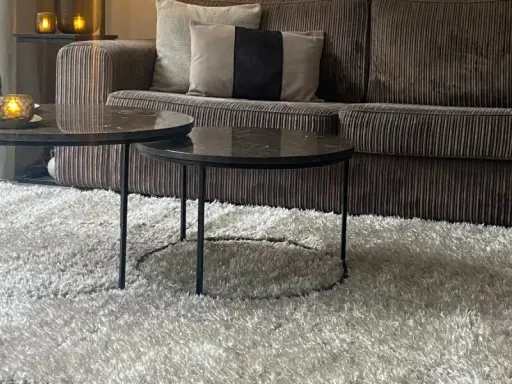 Miękki dywan w kolorze greige otula salon. Na nim dwa stoliki kawowe z marmurowymi blatami i aksamitna kanapa.