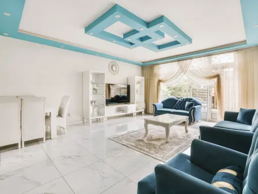Nowoczesny salon z błękitnym, geometrycznym, dekoracyjnym malowidłem na suficie. Wygodne, granatowe sofy i białe meble tworzą elegancką przestrzeń.