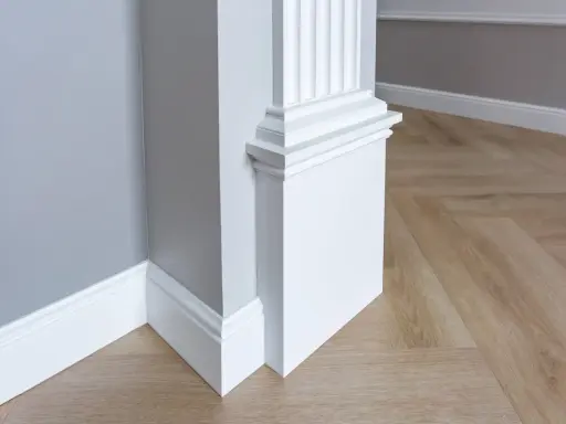 Biały pilaster z żłobieniami i elegancką bazą, idealny do wnętrzarskiego bloga. Drewniana podłoga w jodełkę i szara ściana tworzą stylowe tło.