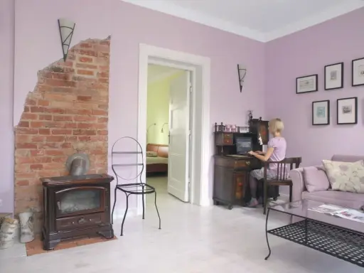 Pokój z ceglanym piecem i biurkiem, gdzie pracuje kobieta. Ściany w odcieniu **kolor lawendowy**.