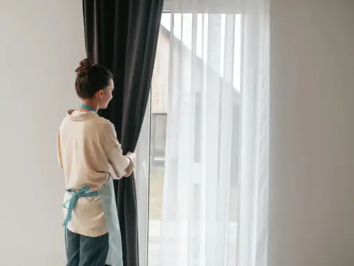 Kobieta w fartuszku zasłania okno ciemną zasłoną. Widać białe firanki i karnisz z wieloma ząbkami.