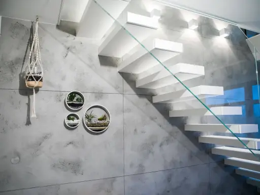 Nowoczesna klatka schodowa z białymi, lewitującymi stopniami i szklaną balustradą. Ściana ozdobiona betonem architektonicznym i wiszącymi doniczkami z sukulentami.
