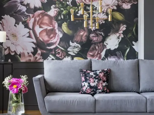 Szara sofa z poduszką w kwiaty, na tle bogatej fototapety. Idealna, by stworzyć przytulną atmosferę.